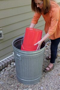 woman placing red plastic 5 gallon bucket upside down in to metal trash can